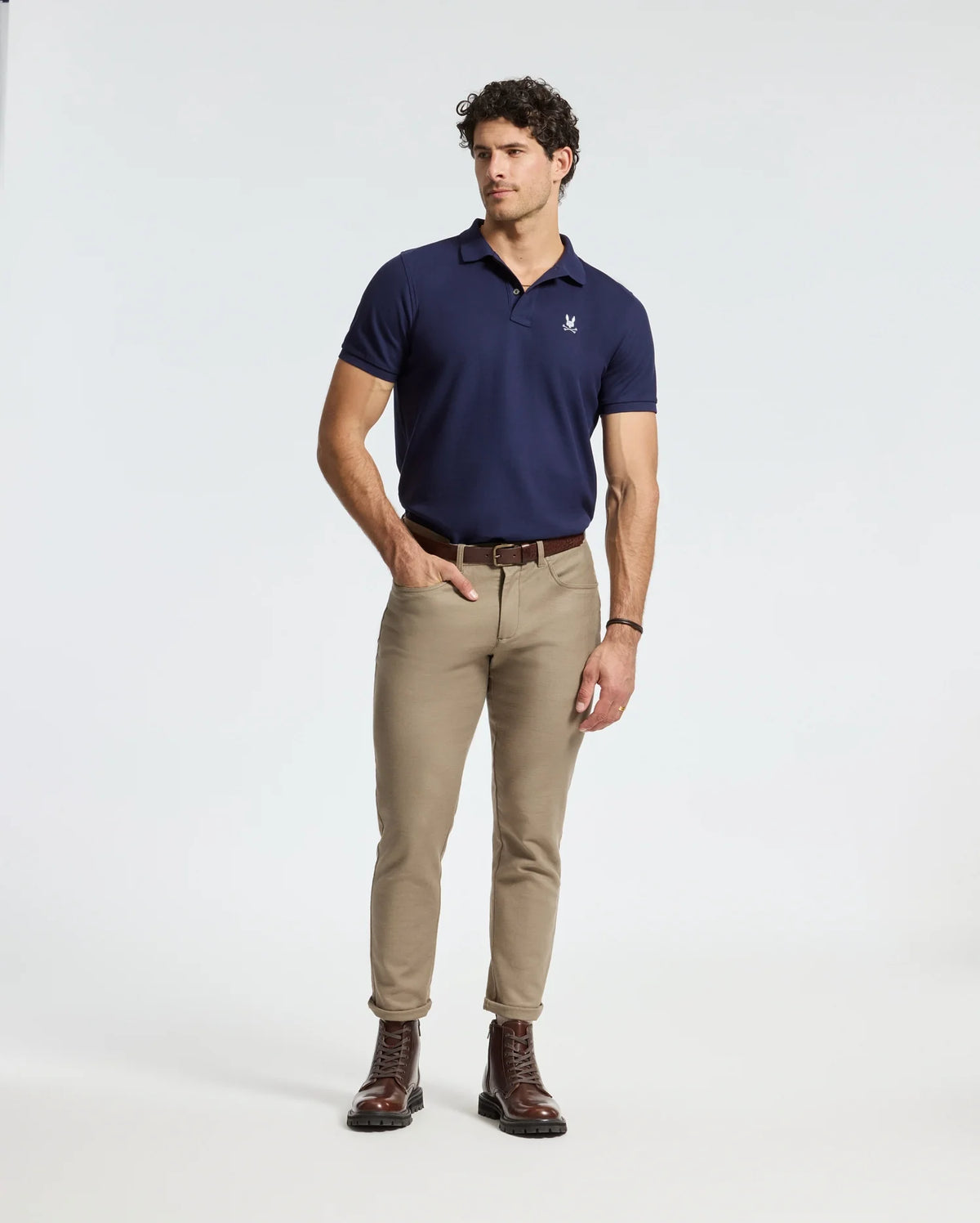 A man with curly hair stands confidently looking to the side, wearing a plush Psycho Bunny MENS ALEXANDER PIQUE POLO and beige pants. His brown boots contribute a rugged touch. With his right hand in his pocket and left hand resting by his side against the plain white background, he exudes effortless style.