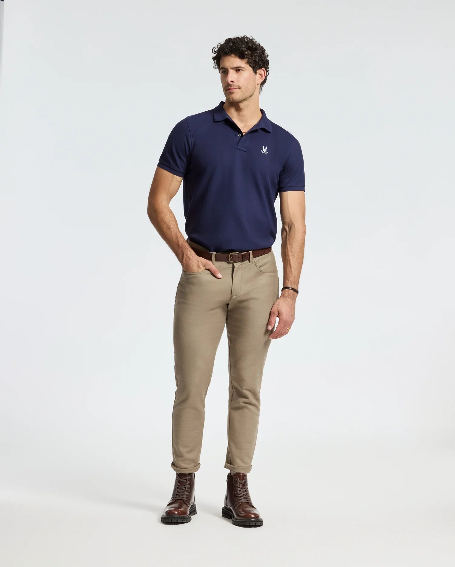A man with curly hair stands confidently looking to the side, wearing a plush Psycho Bunny MENS ALEXANDER PIQUE POLO and beige pants. His brown boots contribute a rugged touch. With his right hand in his pocket and left hand resting by his side against the plain white background, he exudes effortless style.