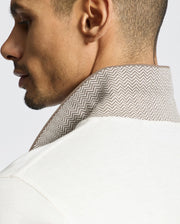 A close-up shows a person in profile wearing the Psycho Bunny MENS ELDORADO PIQUE POLO - B6K105E200, a regular fit white shirt with a textured beige collar made of luxurious Peruvian Pima cotton featuring an elegant herringbone pattern, set against a plain background.