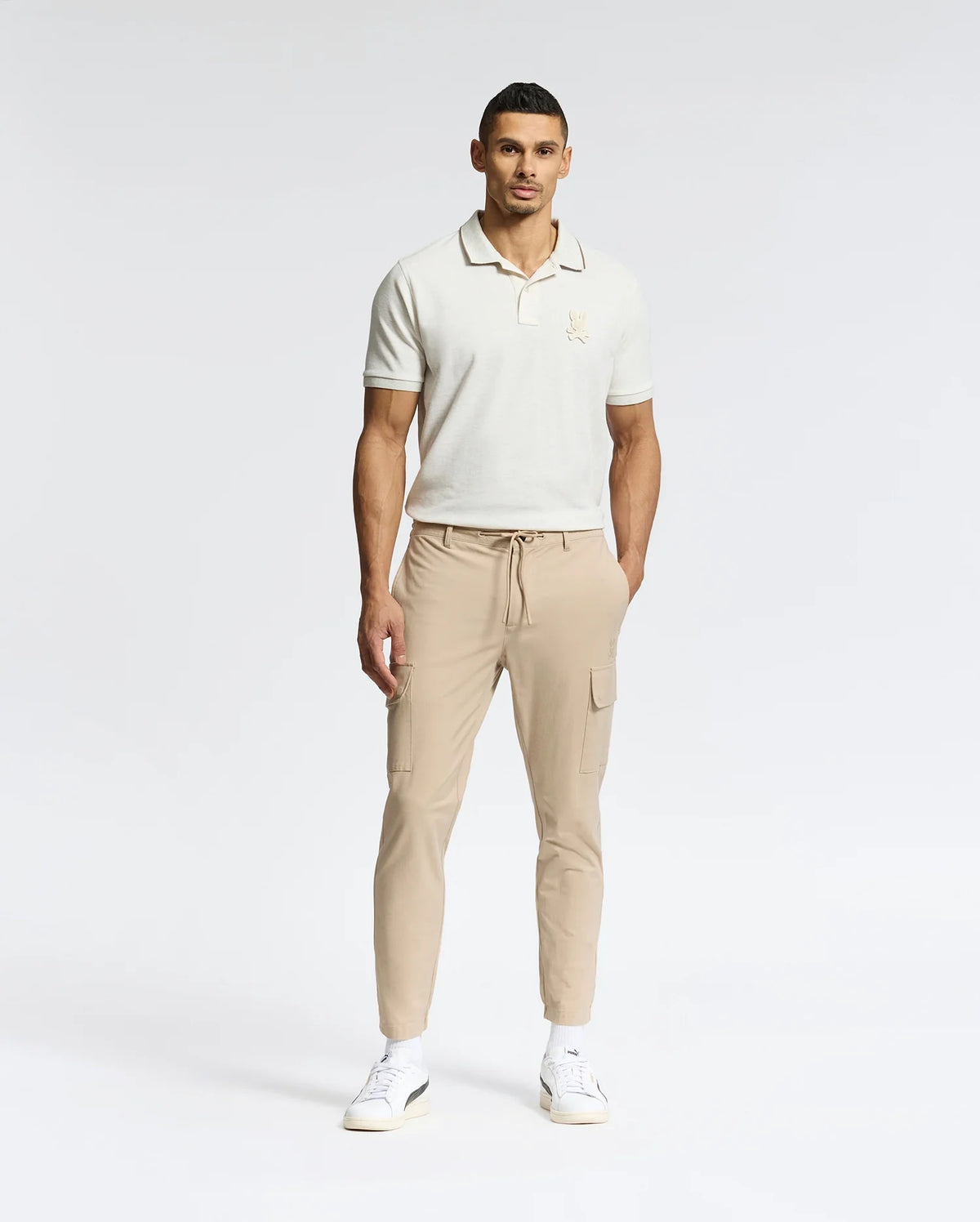 A man, wearing the Psycho Bunny MENS ELDORADO PIQUE POLO (B6K105E200), beige cargo pants, and white sneakers, stands against a plain backdrop. With one hand in his pocket, he gazes neutrally at the camera, highlighting the regular fit attire.