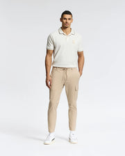 A man, wearing the Psycho Bunny MENS ELDORADO PIQUE POLO (B6K105E200), beige cargo pants, and white sneakers, stands against a plain backdrop. With one hand in his pocket, he gazes neutrally at the camera, highlighting the regular fit attire.