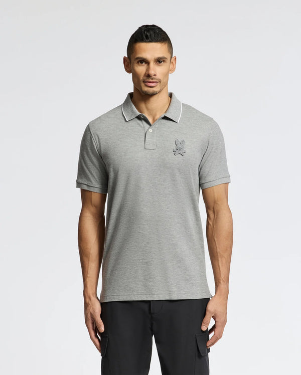 A man in a Psycho Bunny MENS ELDORADO PIQUE POLO (gray Peruvian Pima cotton, with a chenille bunny logo) and black pants poses against a plain white background.