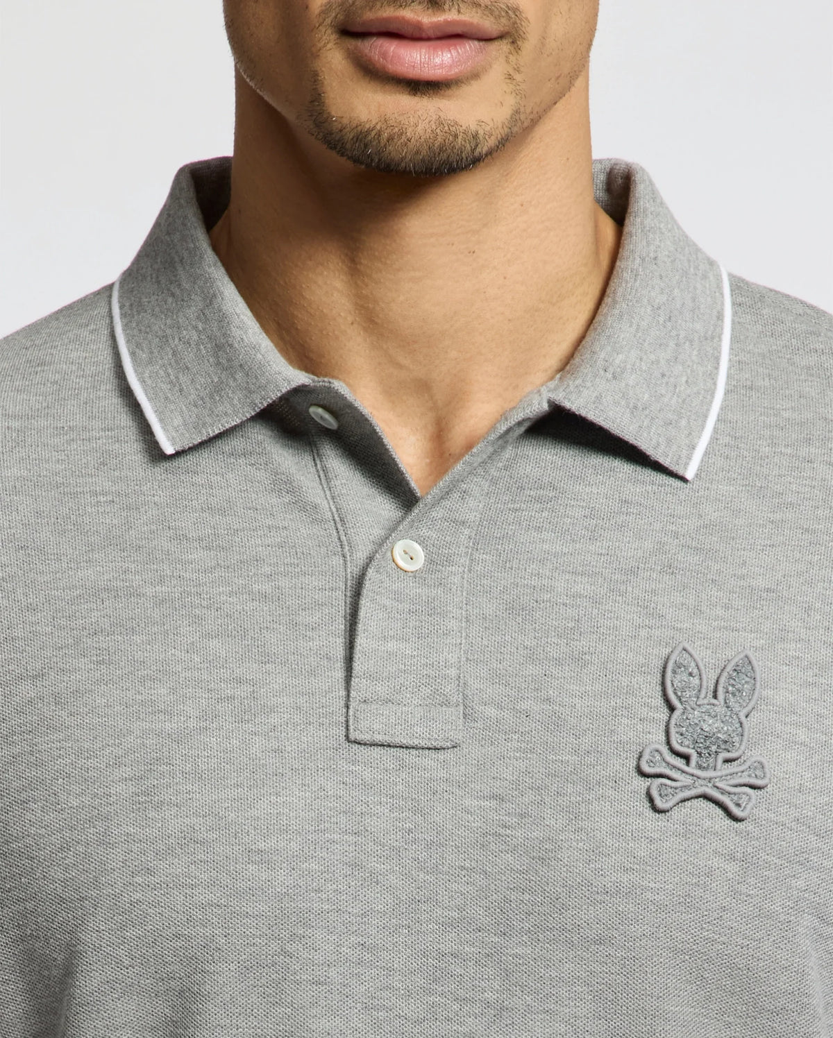 A man wears a Psycho Bunny MENS ELDORADO PIQUE POLO (B6K105E200), crafted from soft Peruvian Pima cotton in gray, with a contrasting white collar trim. Two buttons are fastened, and an embroidered chenille bunny logo is visible. The man's face is partially shown.