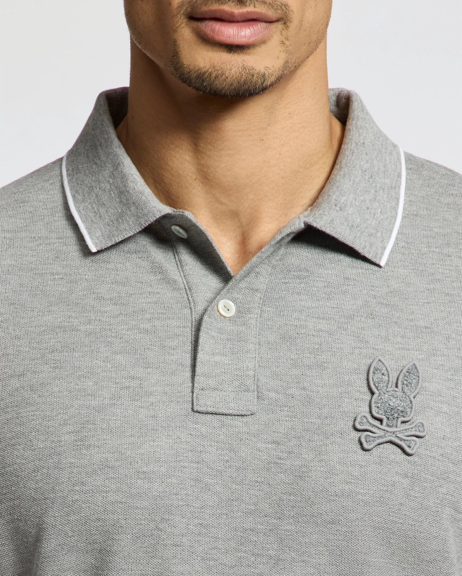 A man wears a Psycho Bunny MENS ELDORADO PIQUE POLO (B6K105E200), crafted from soft Peruvian Pima cotton in gray, with a contrasting white collar trim. Two buttons are fastened, and an embroidered chenille bunny logo is visible. The man's face is partially shown.