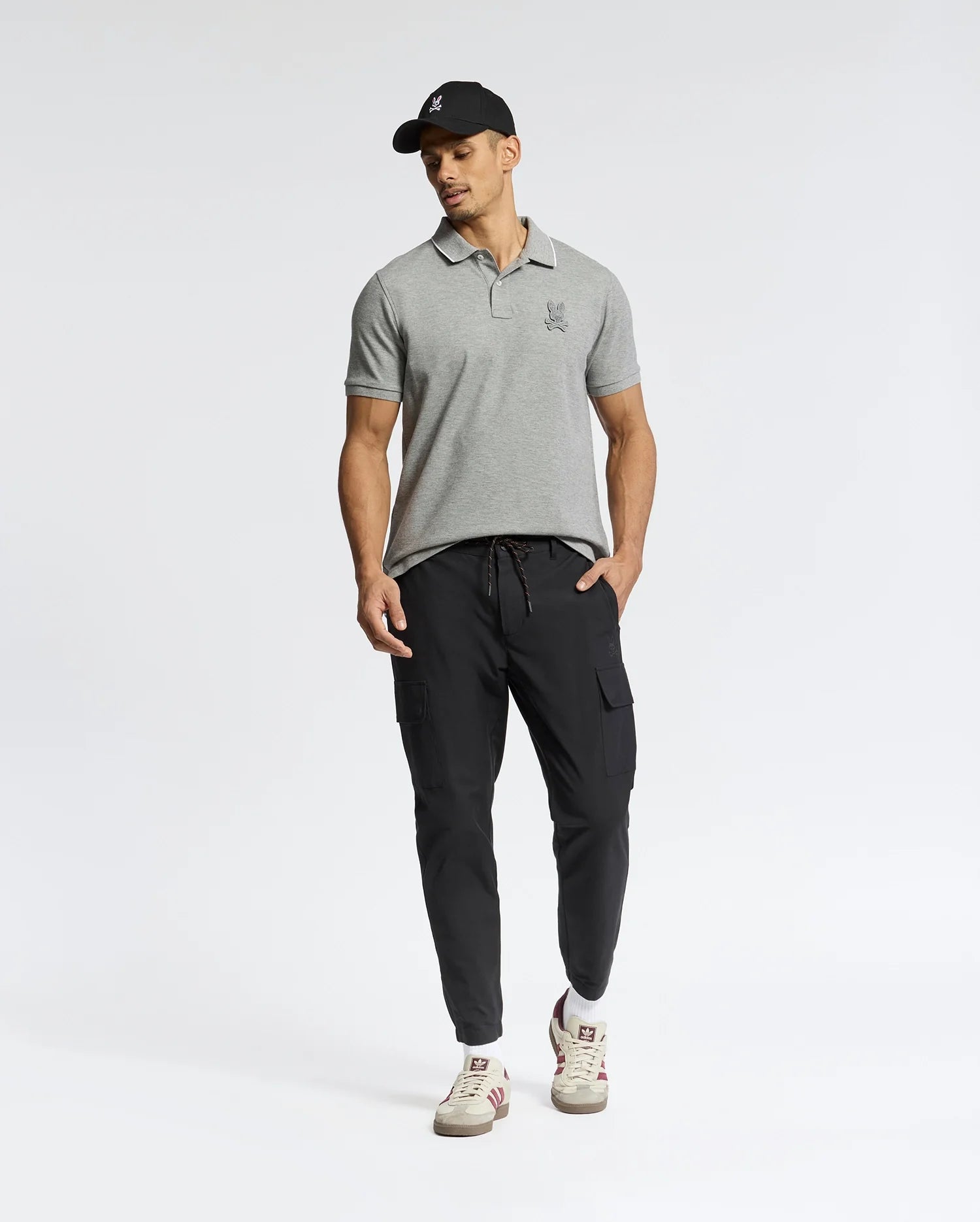 A man wearing a Psycho Bunny Men's Eldorado Pique Polo (B6K105E200), black cargo joggers, white sneakers, and a black cap poses against a plain background.