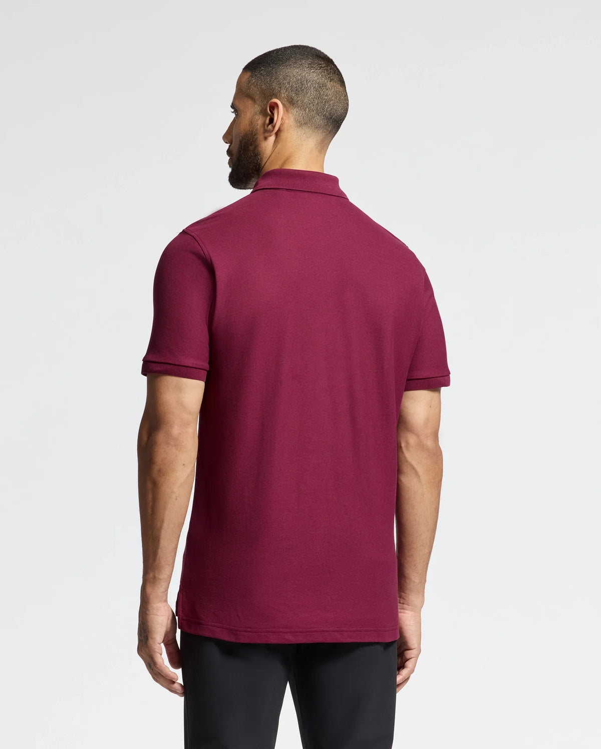 A man with short hair faces away from the camera, dressed in a luxurious Psycho Bunny MENS BRANSON PIQUE POLO - B6K108E200 in maroon, paired with black pants against a plain white background.