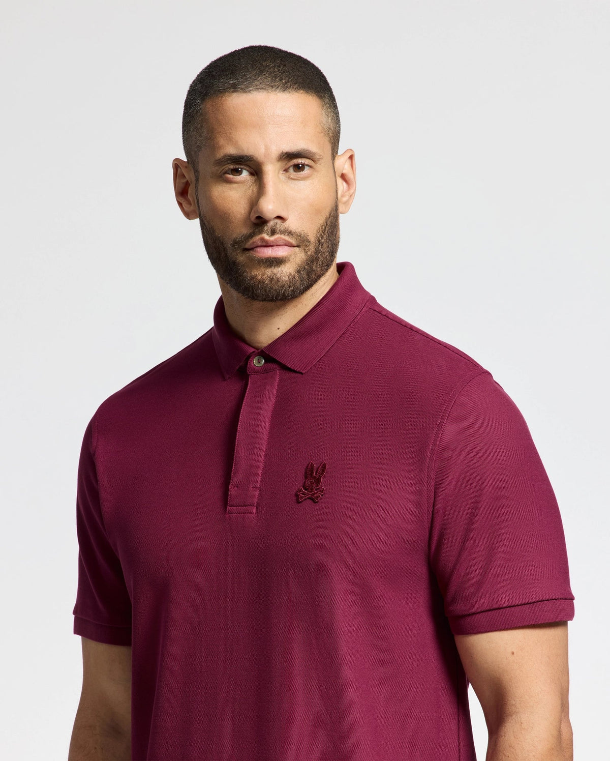 A man with short hair and a beard is dressed in the MENS BRANSON PIQUE POLO - B6K108E200 by Psycho Bunny, featuring an embroidered logo. Crafted from luxurious Pima cotton, this maroon classic men's polo shirt stands out against a plain white background, radiating elegance and timeless style.