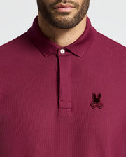 A person wearing a maroon Psycho Bunny Men's Branson Pique Polo (B6K108E200) with an embroidered rabbit head and crossed bones design on the left chest, and the shirt collar buttoned up.