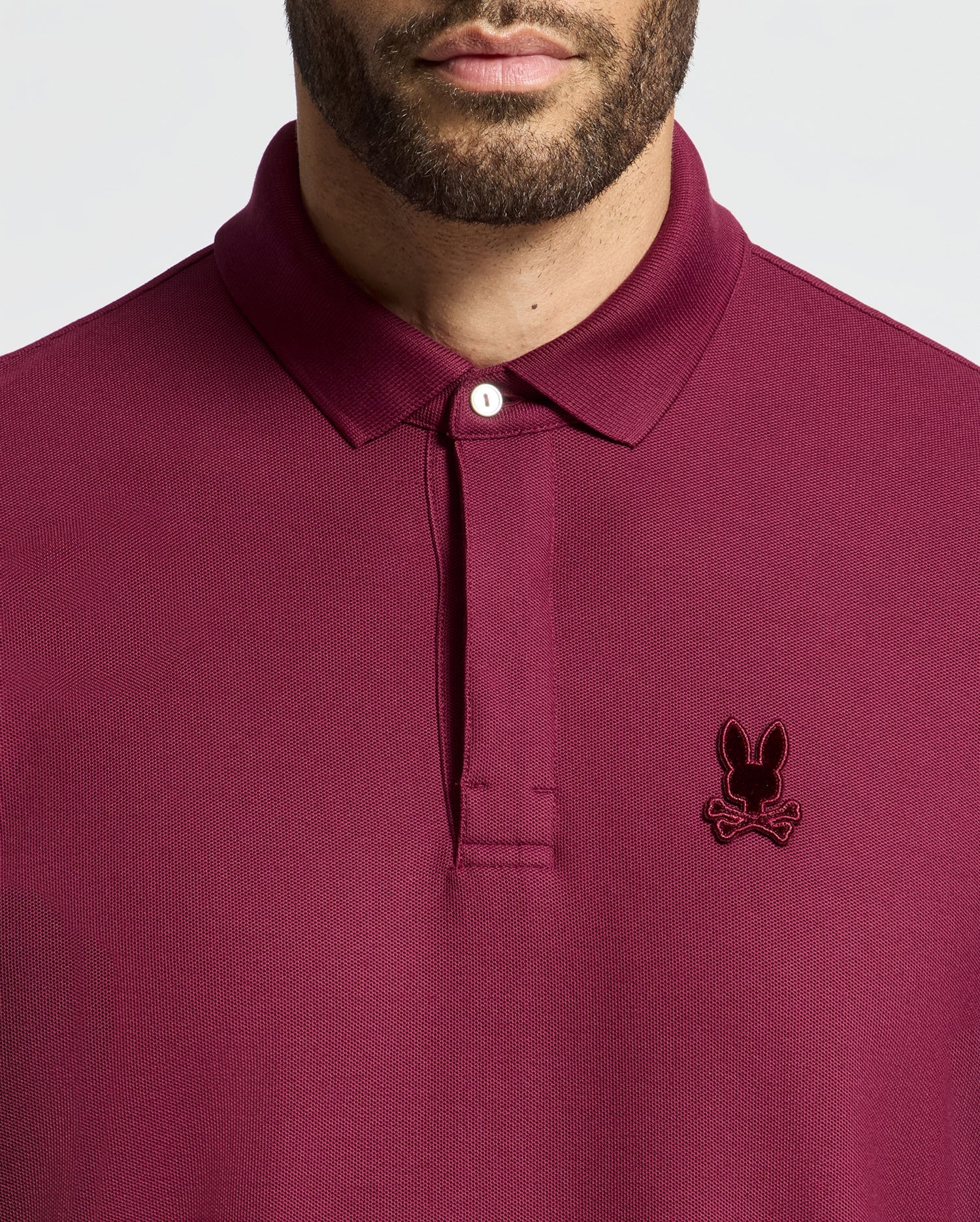 A person wearing a maroon Psycho Bunny Men's Branson Pique Polo (B6K108E200) with an embroidered rabbit head and crossed bones design on the left chest, and the shirt collar buttoned up.