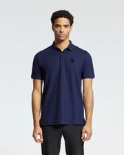 A man stands against a plain background wearing the MENS BRANSON PIQUE POLO - B6K108E200 from Psycho Bunny, featuring a velvet patch logo embroidered on the chest, paired with black pants. He has short, curly hair and maintains a neutral expression.