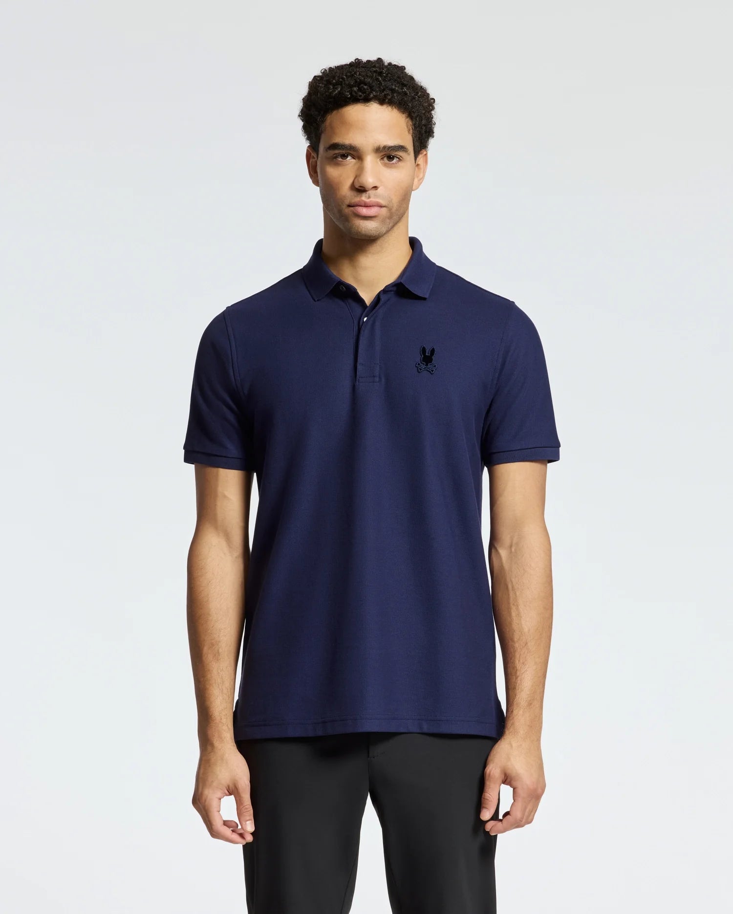A man stands against a plain background wearing the MENS BRANSON PIQUE POLO - B6K108E200 from Psycho Bunny, featuring a velvet patch logo embroidered on the chest, paired with black pants. He has short, curly hair and maintains a neutral expression.
