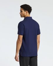 A man with short curly hair is wearing a navy blue MENS BRANSON PIQUE POLO - B6K108E200 by Psycho Bunny and black pants, standing sideways while looking away. The Velvet Patch Logo adds a touch of elegance, set against a plain white background.