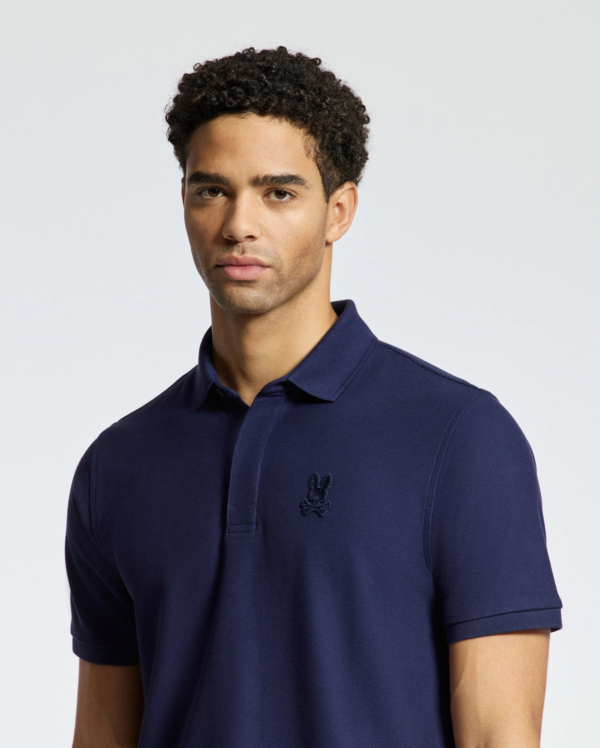 A person with curly hair wearing a navy blue MENS BRANSON PIQUE POLO by Psycho Bunny stands against a plain background.