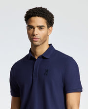 A person with curly hair wearing a navy blue MENS BRANSON PIQUE POLO by Psycho Bunny stands against a plain background.