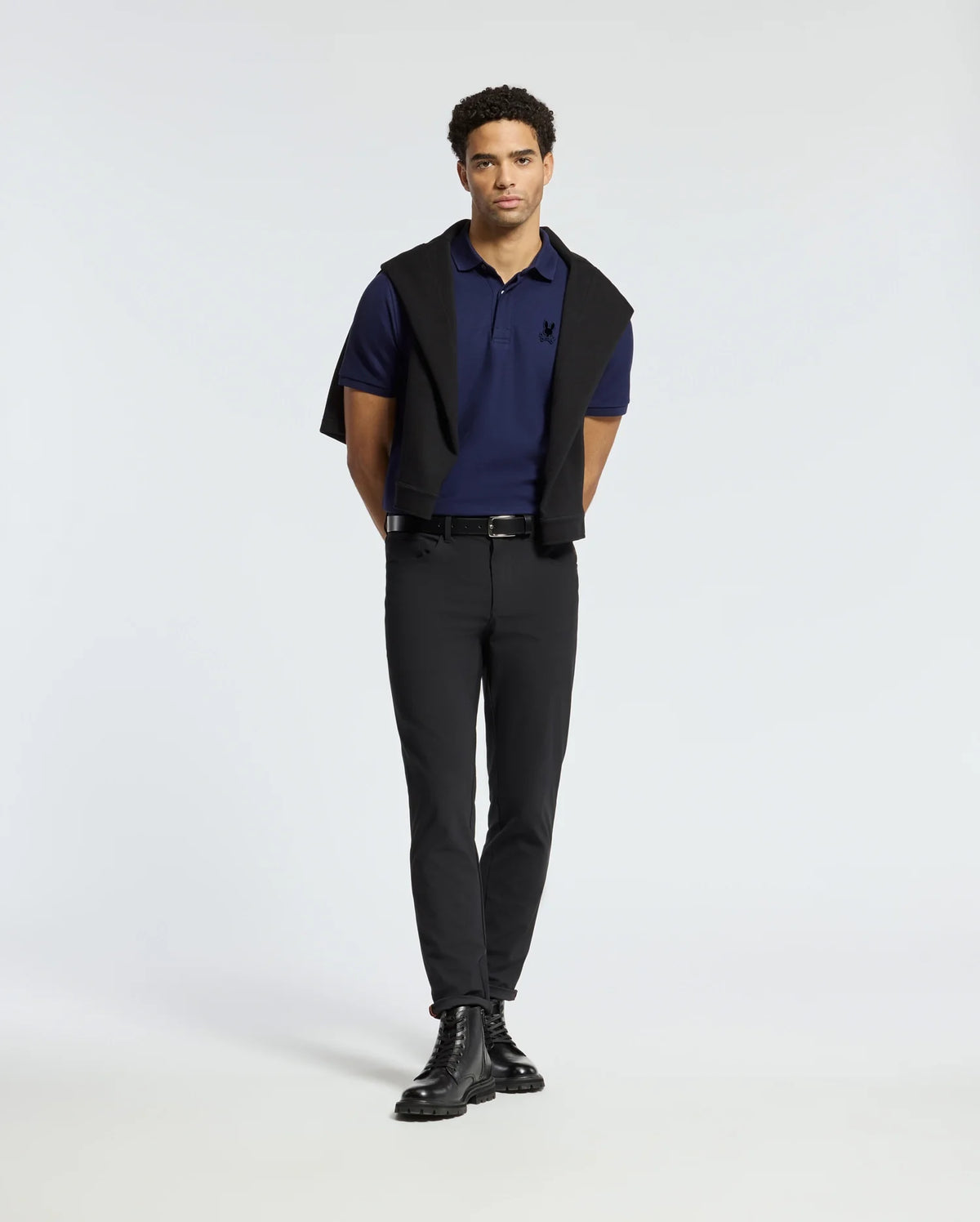 A man stands against a neutral background, showcasing the premium Psycho Bunny Men's Branson Pique Polo in navy, paired stylishly with black pants and boots. A black sweater is elegantly draped over his shoulders with the sleeves tied in front. His short curly hair and direct gaze enhance the sophisticated appearance.
