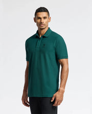 A person wearing a Psycho Bunny MENS BRANSON PIQUE POLO - B6K108E200 in teal, featuring a subtle logo on the chest, stands against a plain white background. The classic polo shirt is paired with dark pants, complementing their short, dark hair as they look directly at the camera.