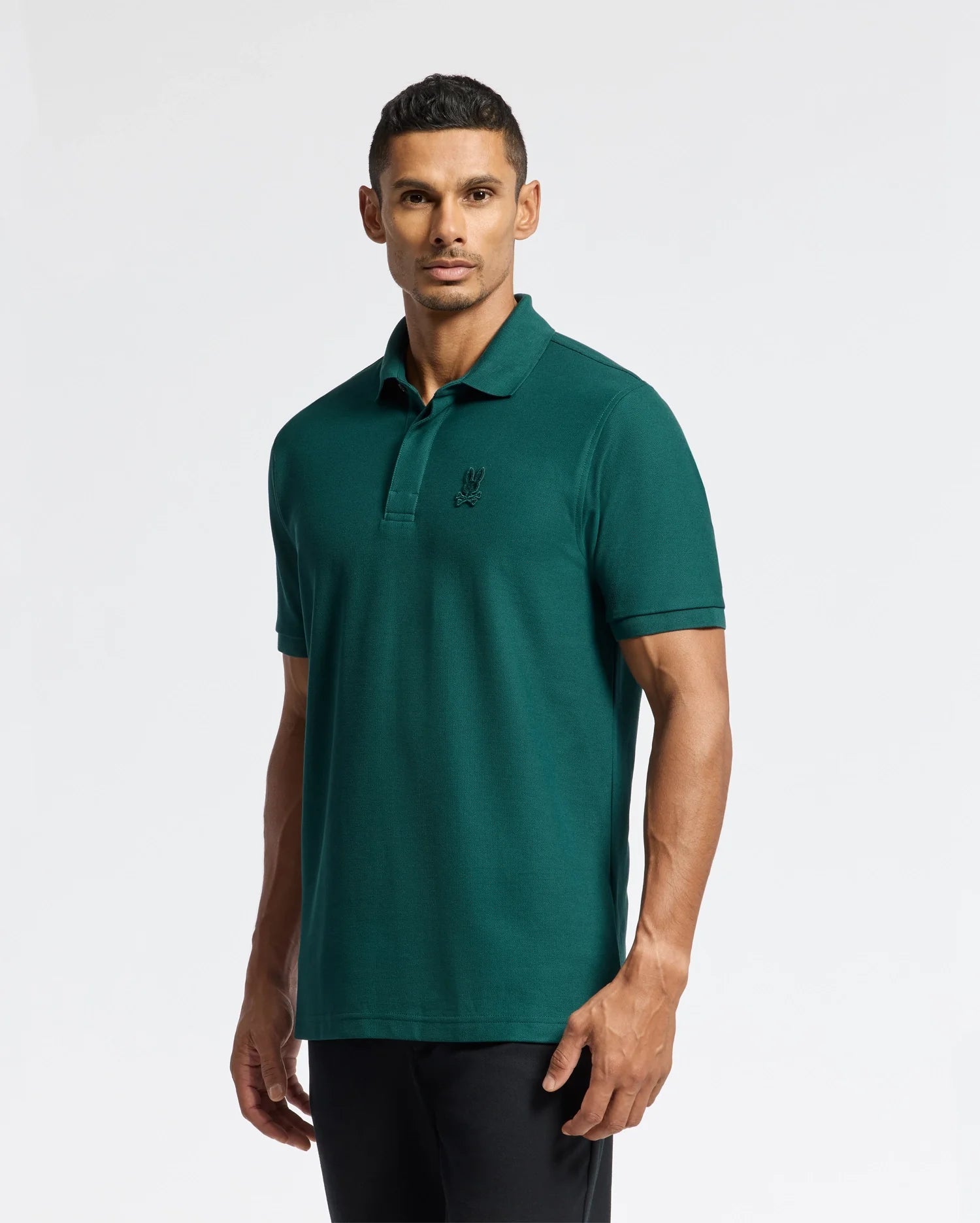 A person wearing a Psycho Bunny MENS BRANSON PIQUE POLO - B6K108E200 in teal, featuring a subtle logo on the chest, stands against a plain white background. The classic polo shirt is paired with dark pants, complementing their short, dark hair as they look directly at the camera.