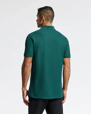 A man with short dark hair stands with his back to the camera, wearing a Psycho Bunny MENS BRANSON PIQUE POLO in dark green and black pants against a plain white background.