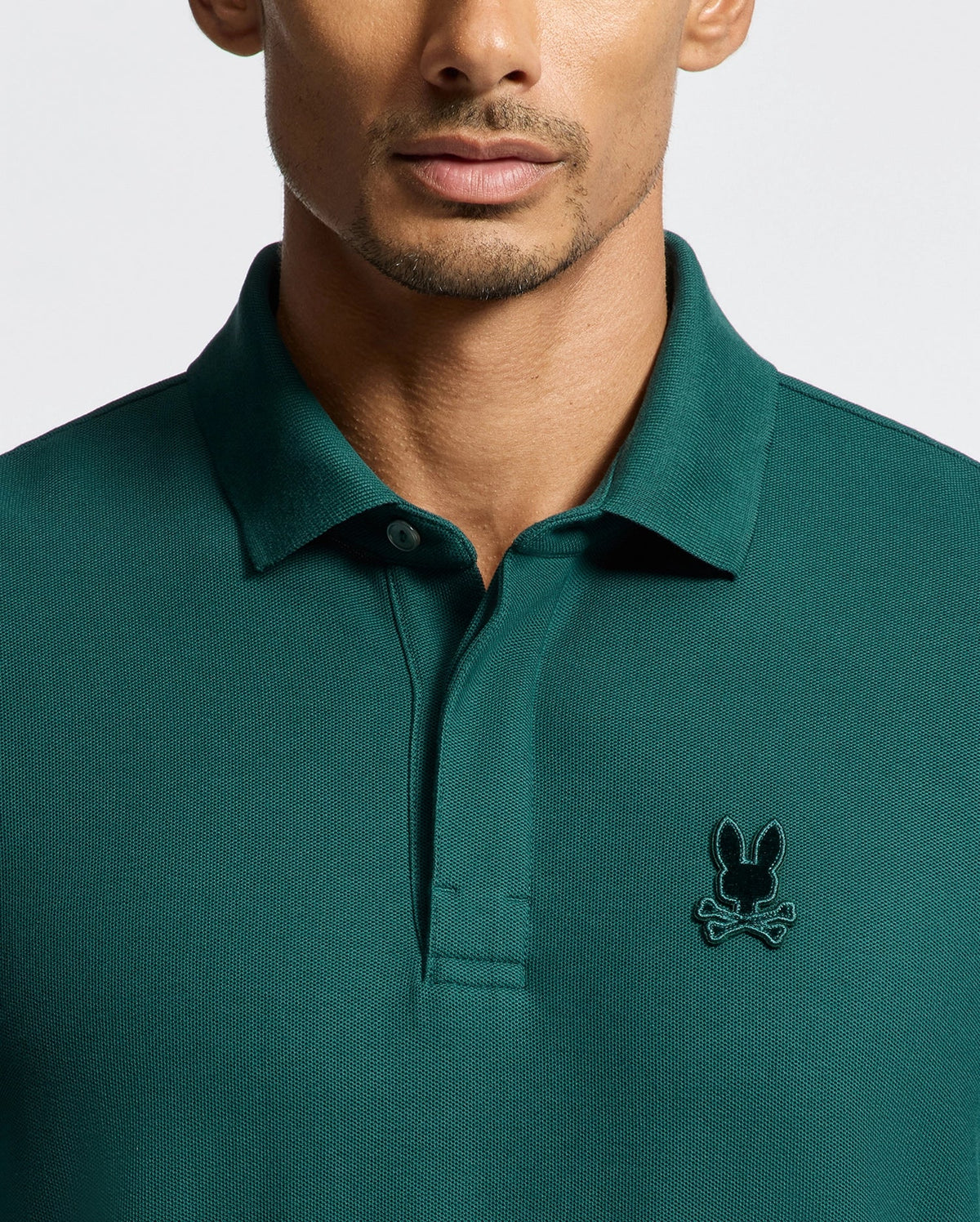A person wearing a Psycho Bunny MENS BRANSON PIQUE POLO in dark green, adorned with a small black rabbit and crossbones logo on the left chest. The plain background accentuates the shirt's detailed embroidery and the person's lower face.