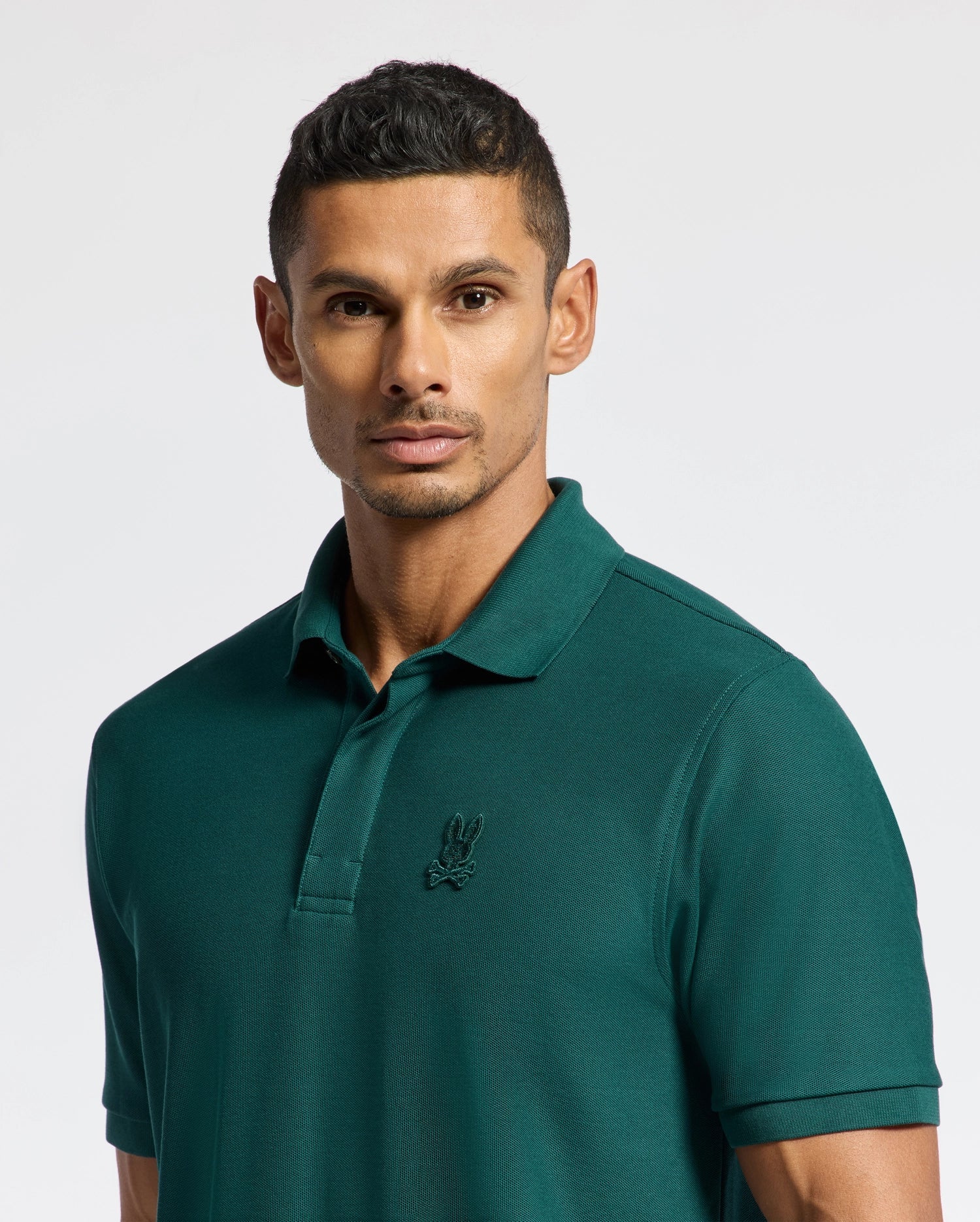 A man with short dark hair is wearing the Psycho Bunny MENS BRANSON PIQUE POLO - B6K108E200, featuring an embroidered logo on the chest. This premium quality polo, in a rich green hue, presents him looking directly at the camera against a light gray background.