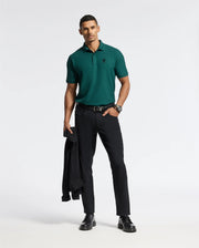 A man stands confidently against a white background, wearing a premium quality Psycho Bunny Men's Branson Pique Polo (B6K108E200) in green, paired with black pants and shoes. He casually holds a black jacket in his left hand while sporting a watch on his wrist.