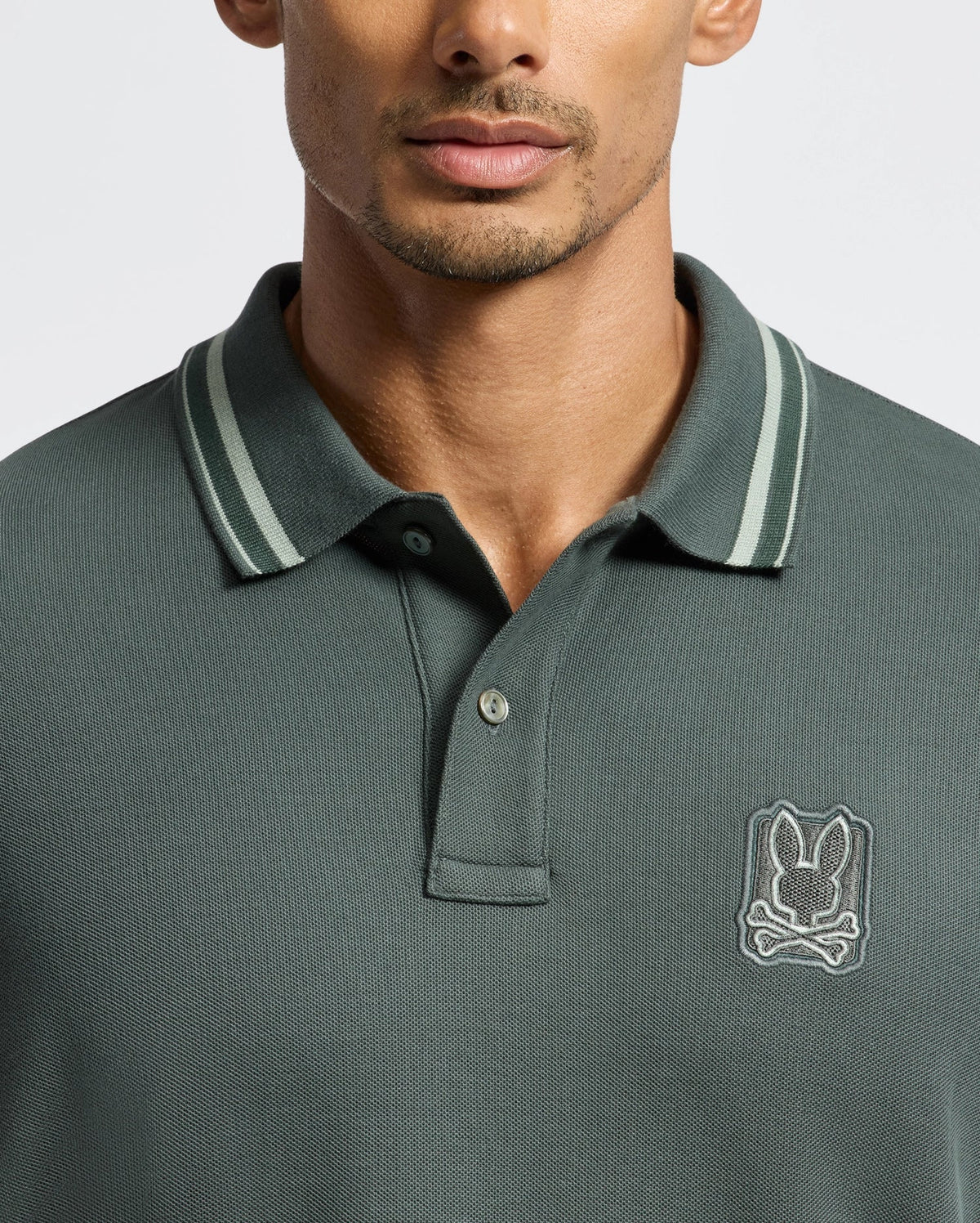 A man is wearing the Psycho Bunny MENS PIERCE PIQUE POLO - B6K201E200 in dark green, featuring a small embroidered logo of a stylized skull and crossbones on the chest. The polo shirt includes a button placket and striped detailing on the collar, set against a plain white background.