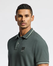 A man with short, dark hair is wearing a dark green MENS PIERCE PIQUE POLO - B6K201E200 from Psycho Bunny, which features an embroidered Bunny logo on the chest. The background is plain white.