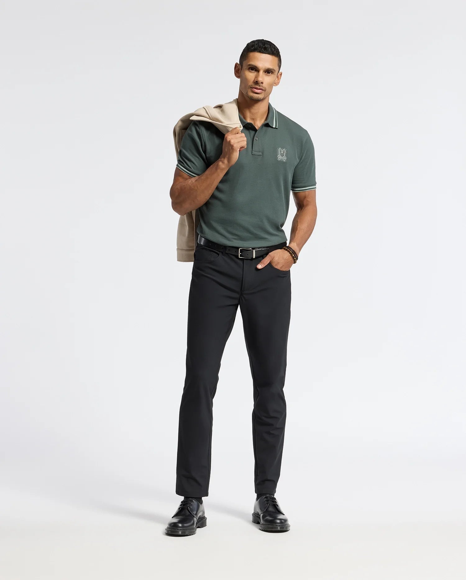 A person stands confidently against a plain background, wearing the dark green MENS PIERCE PIQUE POLO by Psycho Bunny, featuring an embroidered Bunny design. They pair the polo with black pants, hold a light-colored jacket over their shoulder, and complete the look with black shoes.