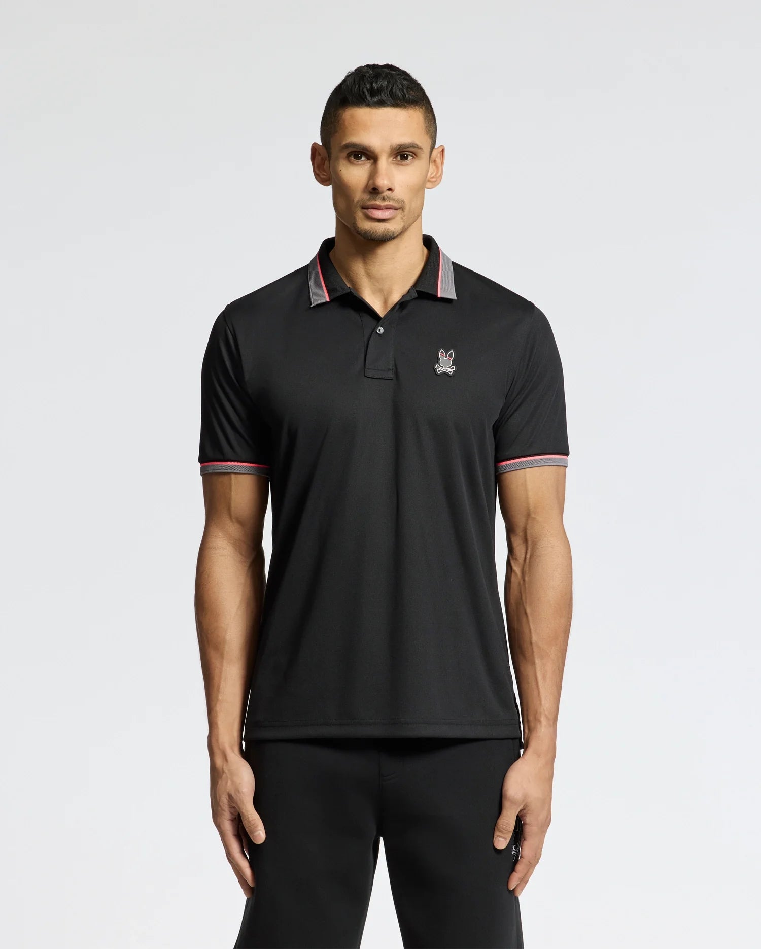 A man stands against a white background wearing the Psycho Bunny MENS STRATTON SPORT PIQUE POLO in black, distinguished by a small emblem on the chest and Peruvian craftsmanship. The shirt is styled with sportswear flair, featuring red and white stripes on the collar and sleeves.