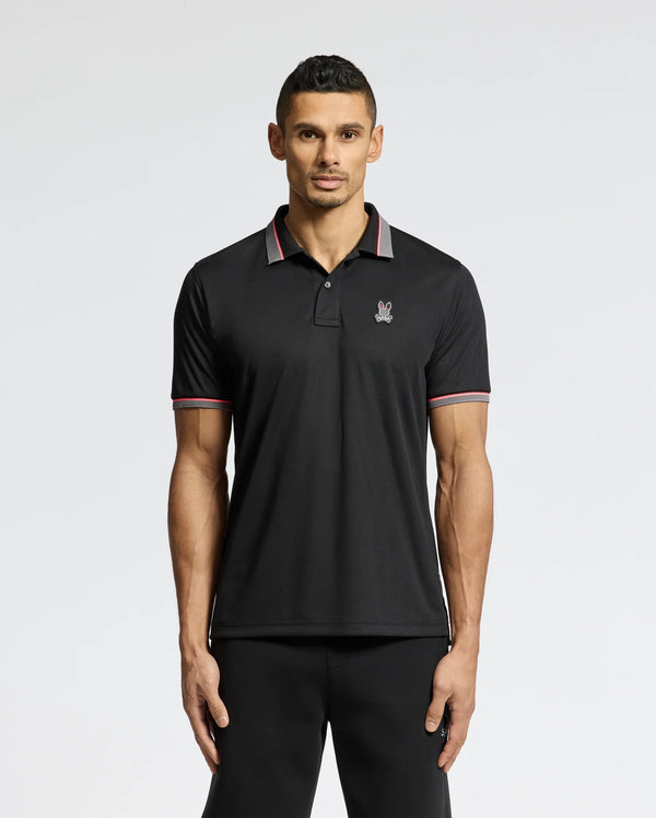 A man stands against a white background wearing the Psycho Bunny MENS STRATTON SPORT PIQUE POLO in black, distinguished by a small emblem on the chest and Peruvian craftsmanship. The shirt is styled with sportswear flair, featuring red and white stripes on the collar and sleeves.