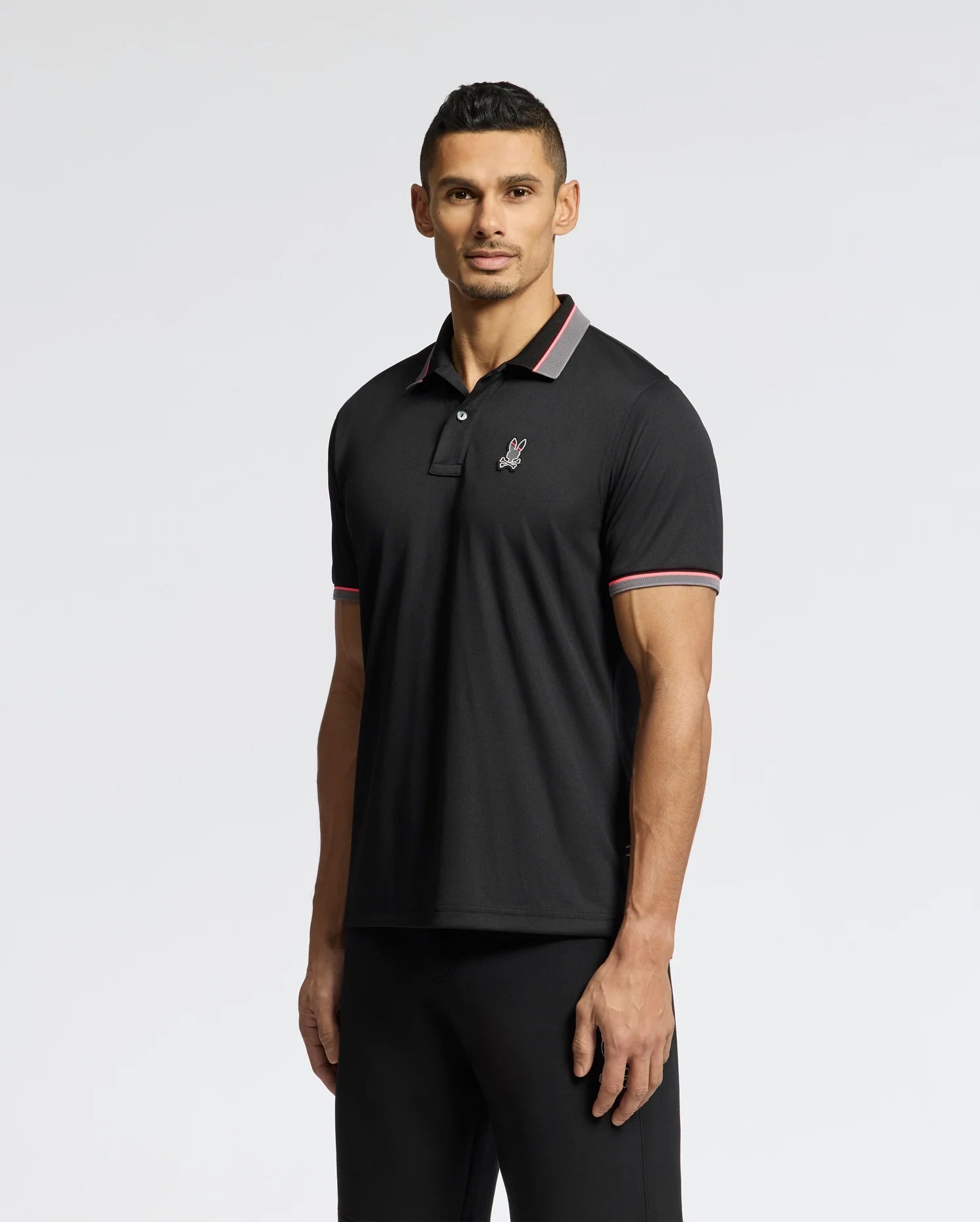 A person stands wearing Psycho Bunny's black Stratton Sport Pique Polo (B6K202E200) and black pants. This Peruvian-crafted polo features a small chest logo and red and white striped collar and sleeve edges, set against a light plain background.