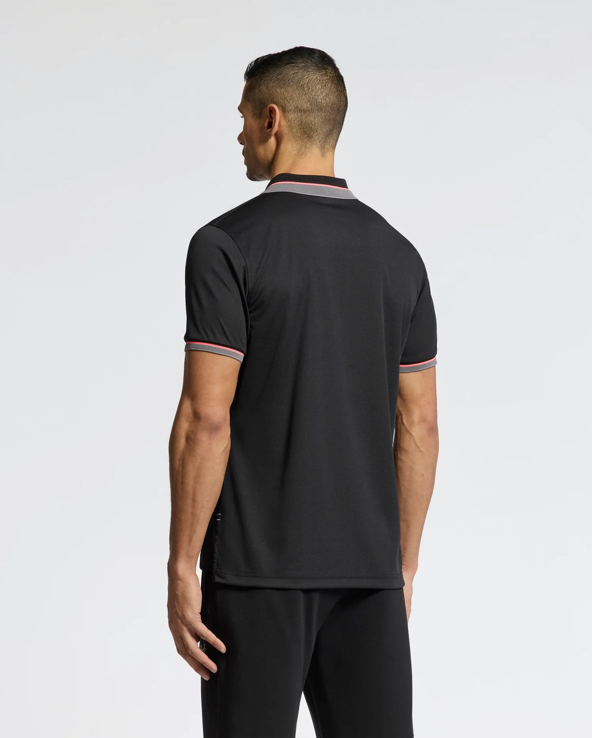 A person with short hair, facing away, wears the Psycho Bunny MENS STRATTON SPORT PIQUE POLO - B6K202E200 in sleek black with red and white trimmed collar and sleeves, paired with black pants. Its moisture-wicking fabric provides comfort against a plain white background.