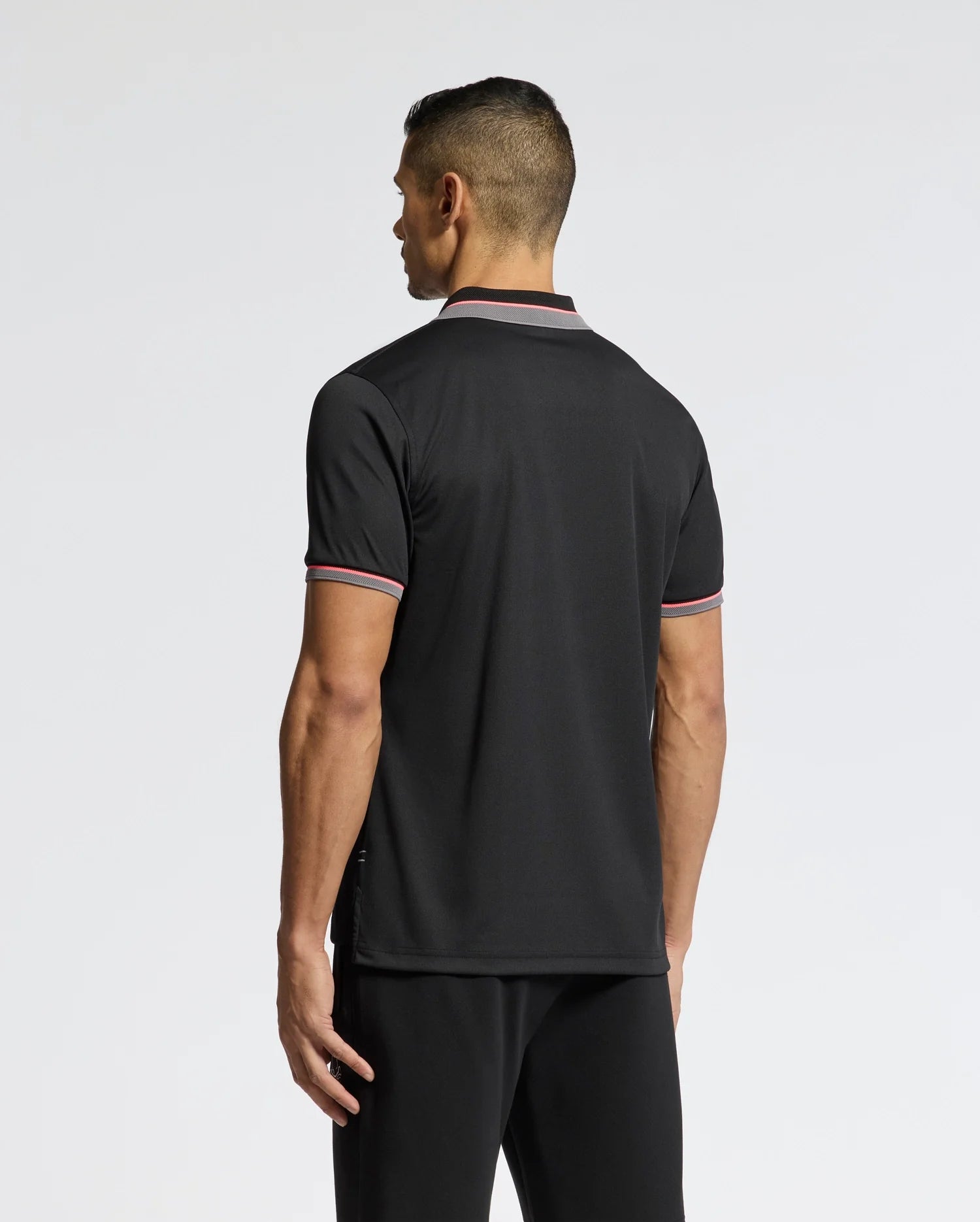 A person with short hair, facing away, wears the Psycho Bunny MENS STRATTON SPORT PIQUE POLO - B6K202E200 in sleek black with red and white trimmed collar and sleeves, paired with black pants. Its moisture-wicking fabric provides comfort against a plain white background.