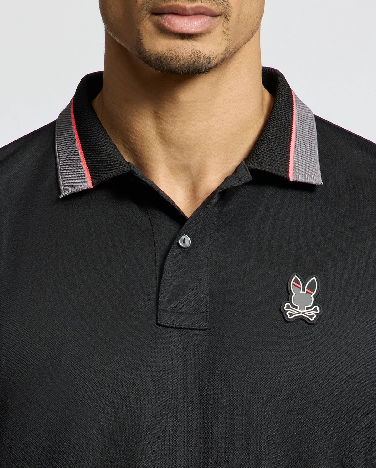 The Psycho Bunny MENS STRATTON SPORT PIQUE POLO - B6K202E200 features a black design with the bunny skull and crossbones logo on the chest. A red and gray two-tone collar adds sporty flair, complemented by moisture-wicking fabric for comfort against a plain white background.
