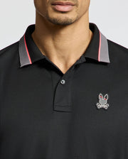 The Psycho Bunny MENS STRATTON SPORT PIQUE POLO - B6K202E200 features a black design with the bunny skull and crossbones logo on the chest. A red and gray two-tone collar adds sporty flair, complemented by moisture-wicking fabric for comfort against a plain white background.