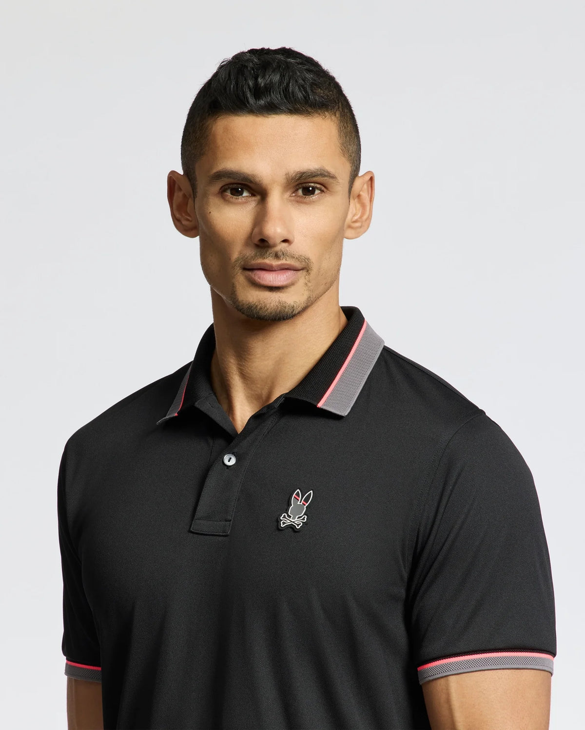 A man wearing a Psycho Bunny MENS STRATTON SPORT PIQUE POLO (B6K202E200) with moisture-wicking fabric and an embroidered rabbit emblem. The black shirt has pink and gray collar accents, showcasing Peruvian craftsmanship. He has short dark hair, a trimmed beard, and stands against a plain light background.