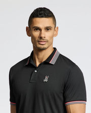 A man wearing a Psycho Bunny MENS STRATTON SPORT PIQUE POLO (B6K202E200) with moisture-wicking fabric and an embroidered rabbit emblem. The black shirt has pink and gray collar accents, showcasing Peruvian craftsmanship. He has short dark hair, a trimmed beard, and stands against a plain light background.