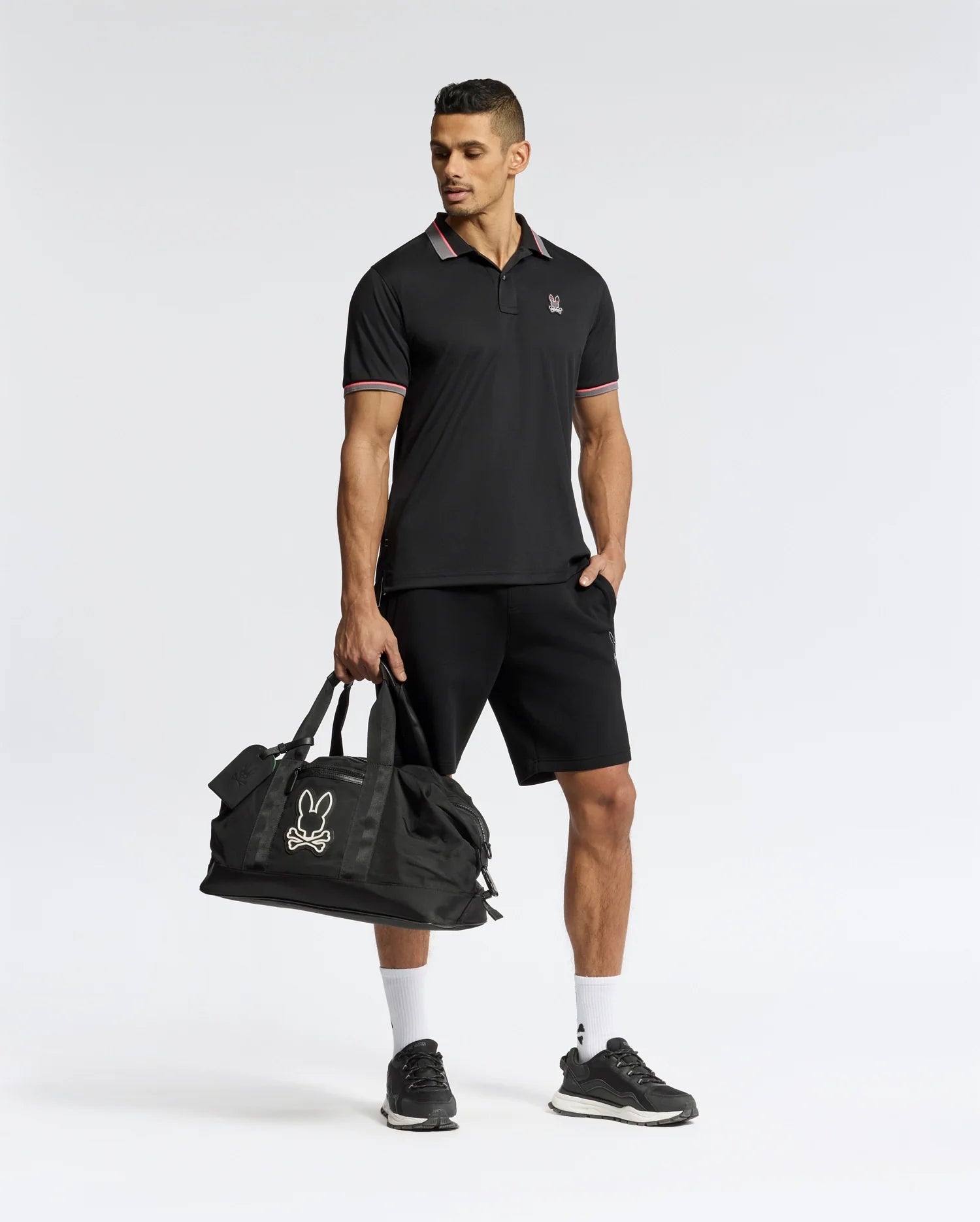 A man in a Psycho Bunny MENS STRATTON SPORT PIQUE POLO, black shorts, and moisture-wicking sneakers holds a large duffel bag with the bunny logo. The plain white background highlights his athletic sportswear.
