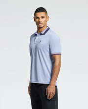 A man in a Psycho Bunny MENS STRATTON SPORT PIQUE POLO (B6K202E200), designed as premium sportswear, stands against a plain background. The light blue shirt boasts moisture-wicking technology, with a small chest logo and dark collar and sleeve edges. He has short hair and looks directly at the camera.