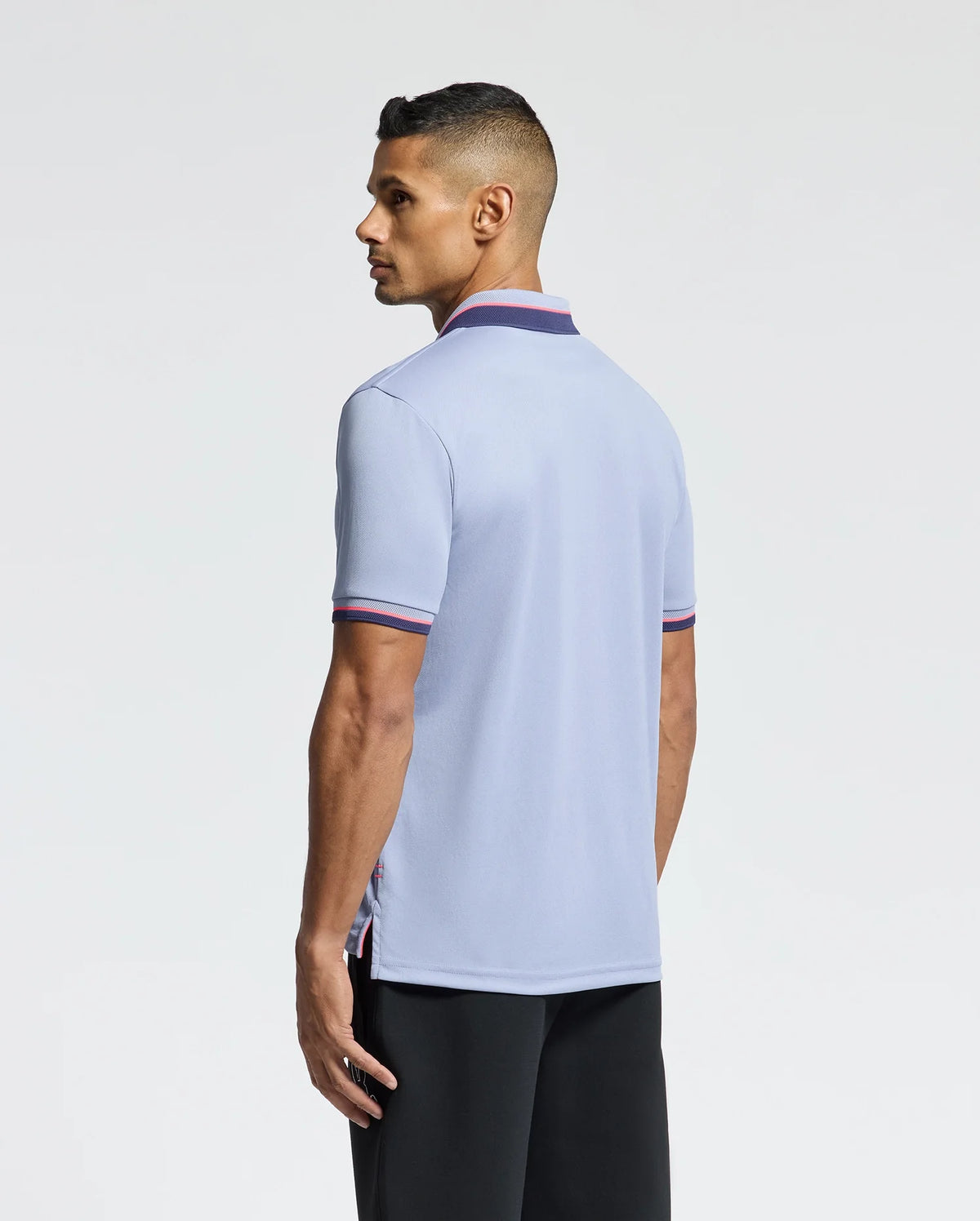 A man with short hair wears the Psycho Bunny MENS STRATTON SPORT PIQUE POLO (B6K202E200) in light blue, featuring contrasting sleeve and collar trim. He faces slightly away from the camera, paired with dark pants against a plain white backdrop, highlighting its sporty style.