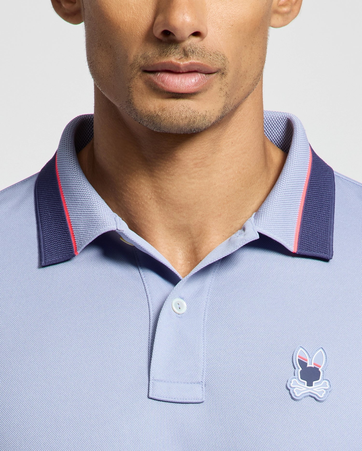 Sporting a Psycho Bunny MENS STRATTON SPORT PIQUE POLO in light blue with a dark blue collar and red trim, this stylish moisture-wicking top, featuring the playful bunny logo on the chest, is perfect for sportswear aficionados. The image focuses on the upper torso to highlight its design.