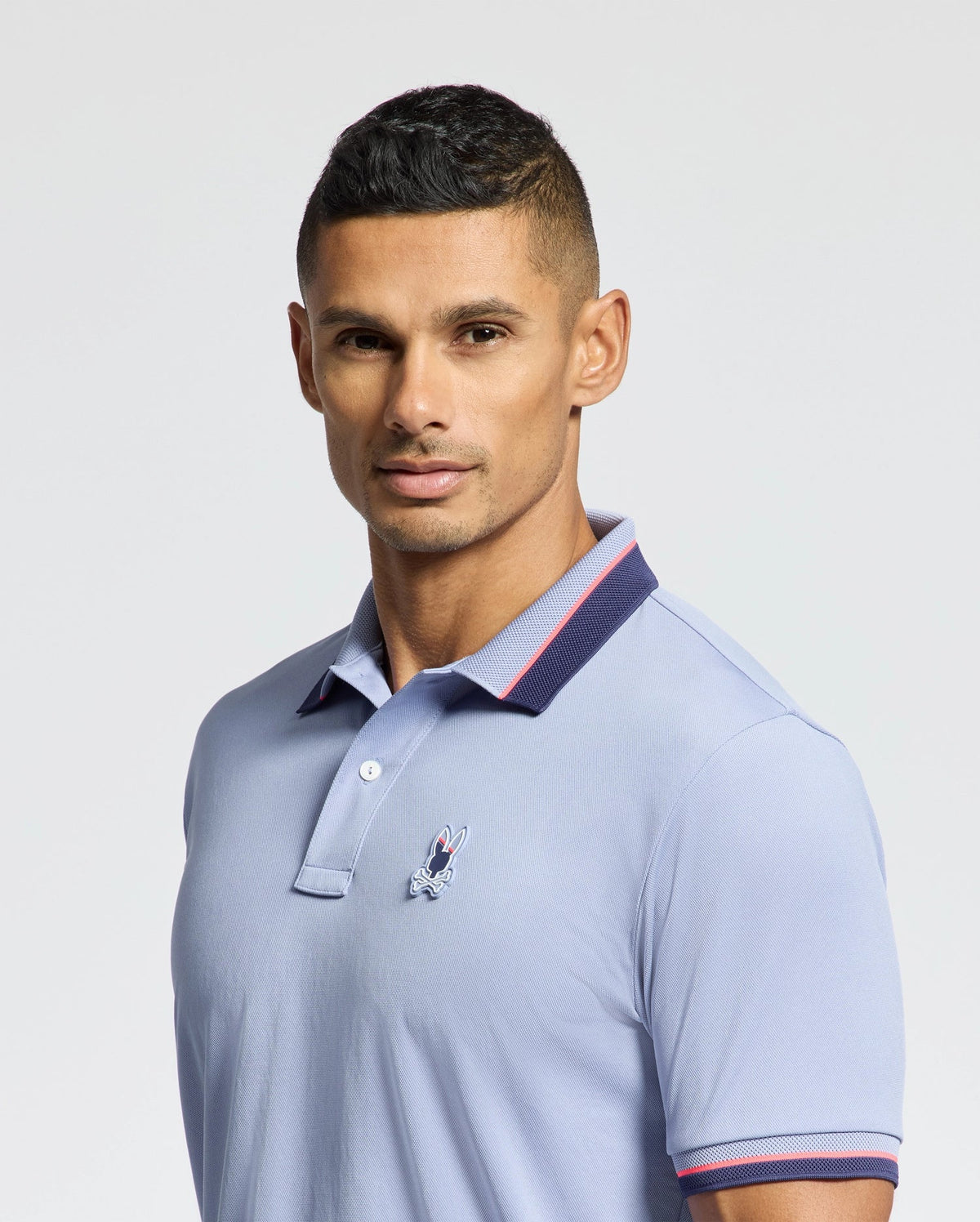 A person wearing the Psycho Bunny MENS STRATTON SPORT PIQUE POLO - B6K202E200, featuring moisture-wicking light blue fabric with a small chest embroidery and a collar with dark trim, poses against a plain background. The individual has short, dark hair and faces the camera directly.