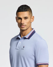 A person wearing the Psycho Bunny MENS STRATTON SPORT PIQUE POLO - B6K202E200, featuring moisture-wicking light blue fabric with a small chest embroidery and a collar with dark trim, poses against a plain background. The individual has short, dark hair and faces the camera directly.