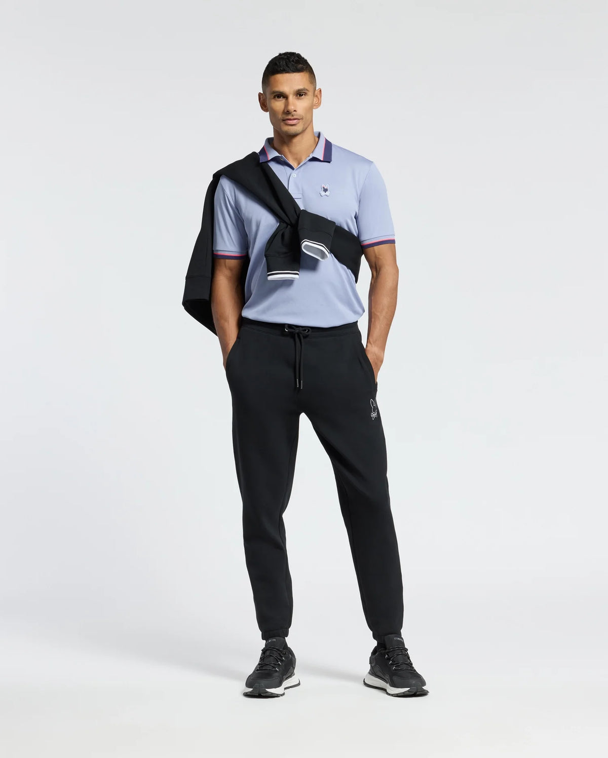 A person exudes confidence wearing the Psycho Bunny MENS STRATTON SPORT PIQUE POLO in light blue, with moisture-wicking technology. It is paired with black joggers and sneakers. A black jacket drapes over their shoulder, completing a stylish sportswear look against a plain white background.