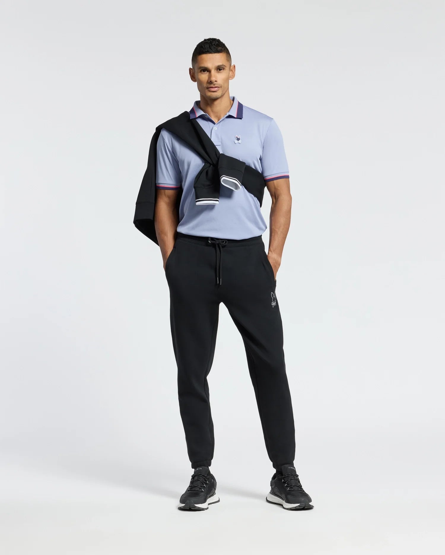 A person exudes confidence wearing the Psycho Bunny MENS STRATTON SPORT PIQUE POLO in light blue, with moisture-wicking technology. It is paired with black joggers and sneakers. A black jacket drapes over their shoulder, completing a stylish sportswear look against a plain white background.