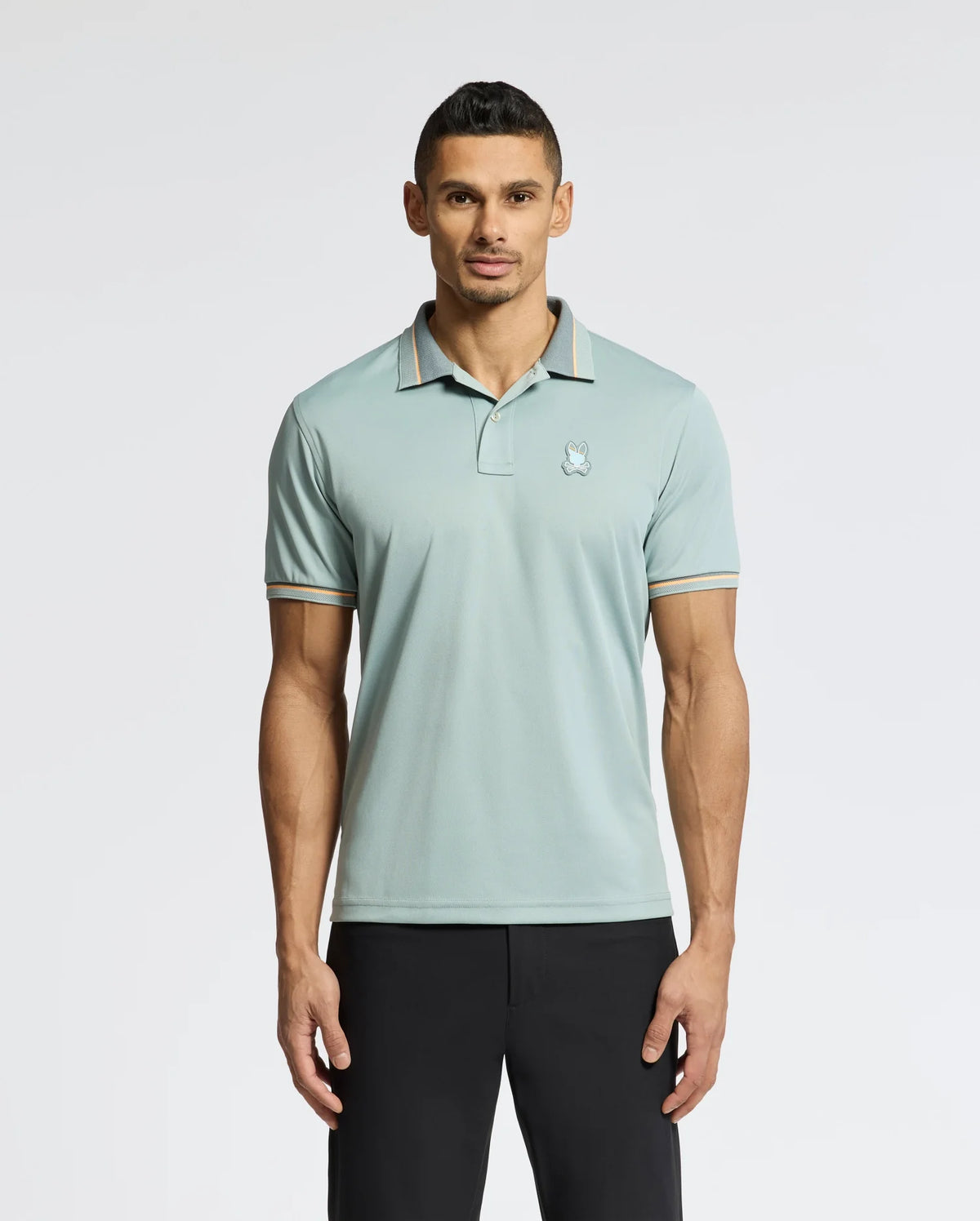 A man with short dark hair stands against a plain background, wearing Psycho Bunny's MENS STRATTON SPORT PIQUE POLO - B6K202E200 in light teal with striped edges and a small chest logo, paired with black pants.