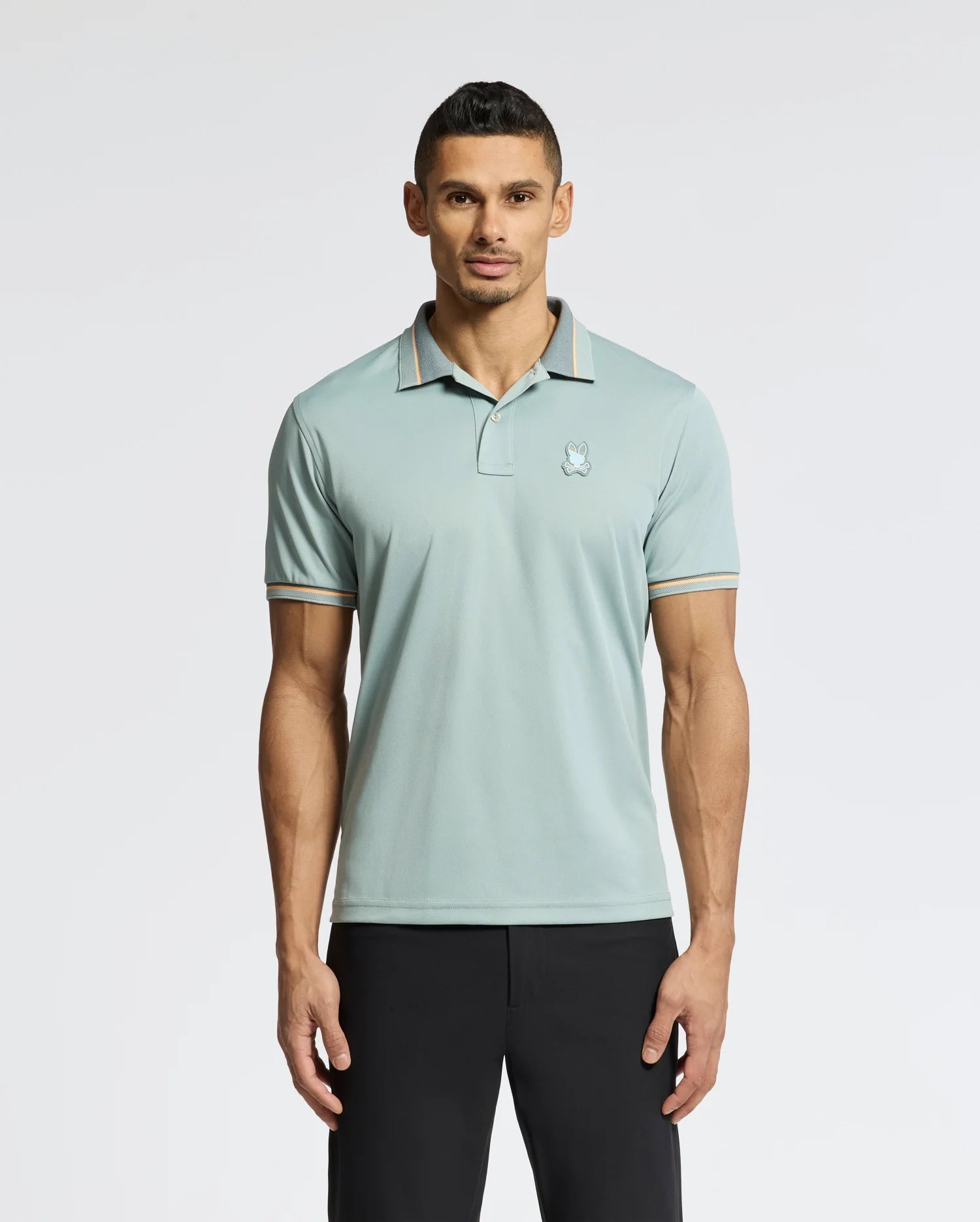 A man with short dark hair stands against a plain background, wearing Psycho Bunny's MENS STRATTON SPORT PIQUE POLO - B6K202E200 in light teal with striped edges and a small chest logo, paired with black pants.