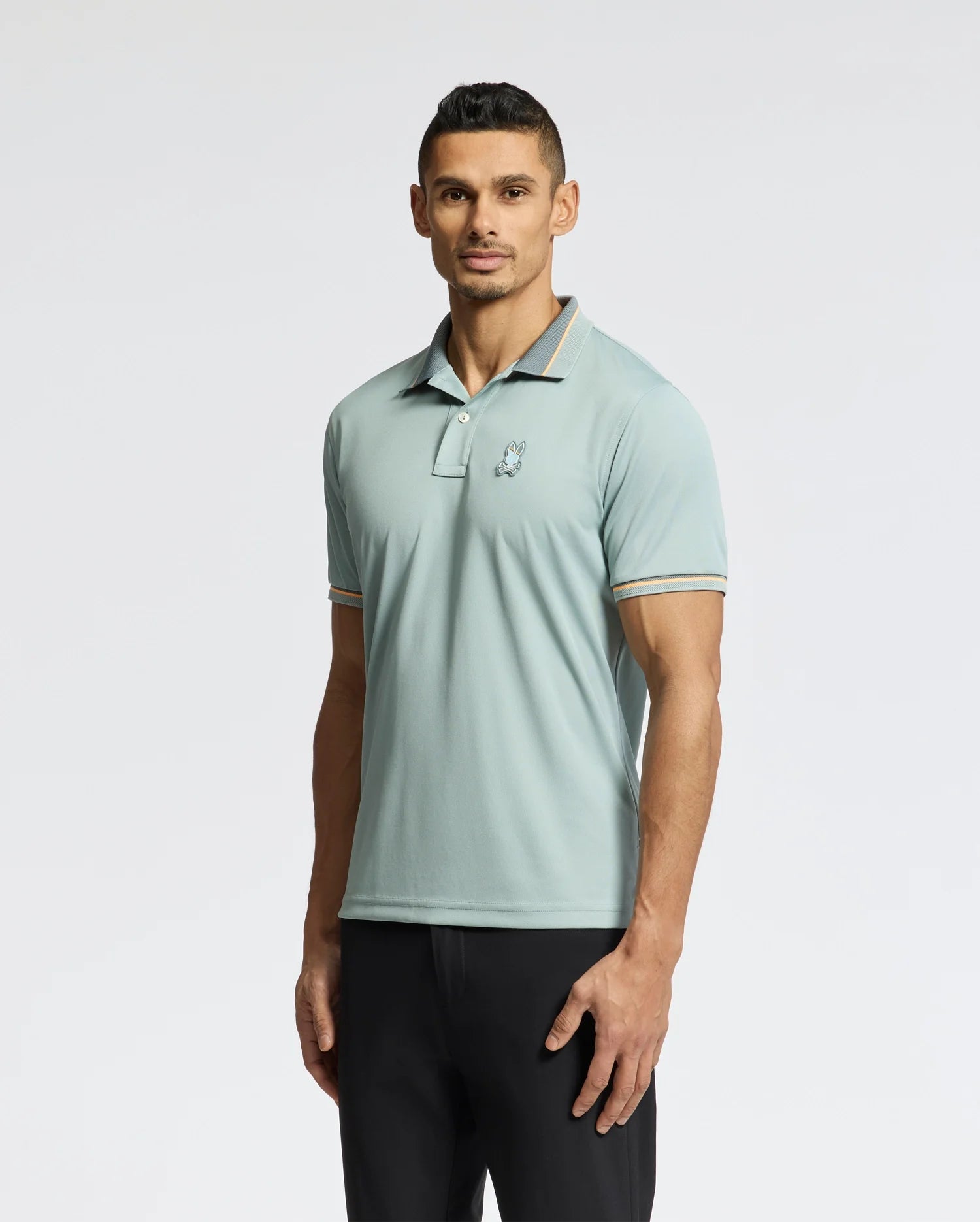 A man in a Psycho Bunny light blue MENS STRATTON SPORT PIQUE POLO - B6K202E200 and dark trousers stands against a white background, facing forward.