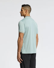 A man with short dark hair stands sideways, facing left, in a Psycho Bunny MENS STRATTON SPORT PIQUE POLO - B6K202E200 in pale blue and black pants, ideal for sportswear lovers. The background is plain white.