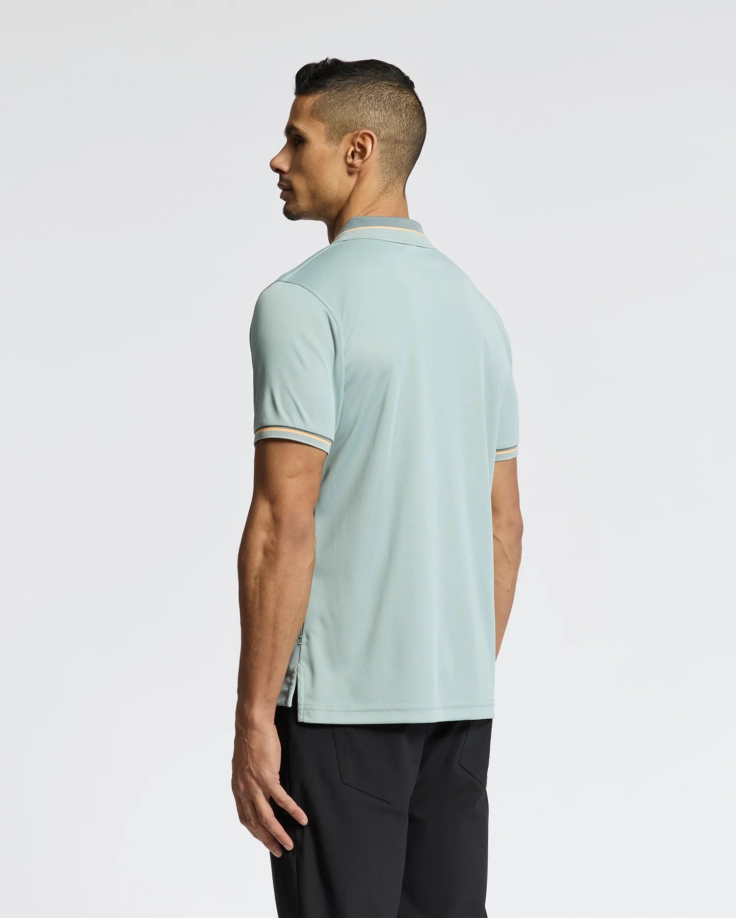 A man with short dark hair stands sideways, facing left, in a Psycho Bunny MENS STRATTON SPORT PIQUE POLO - B6K202E200 in pale blue and black pants, ideal for sportswear lovers. The background is plain white.